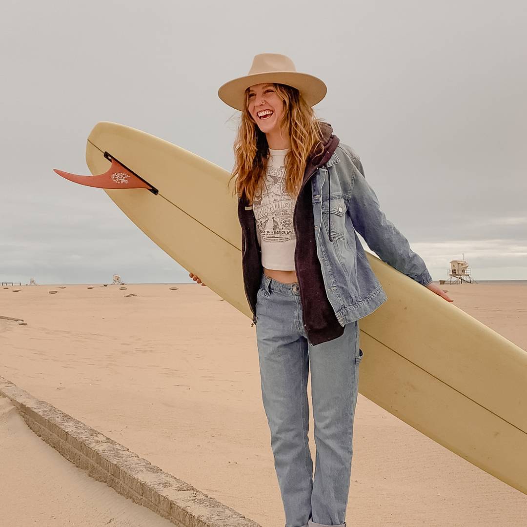 Girl with long hair wearing a wide brim hat and holding a surf board